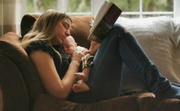 Moment paisible : Marjorie lit un livre sur un canapé, son bébé endormi blotti contre elle, avec une lumière douce filtrant à travers une fenêtre.